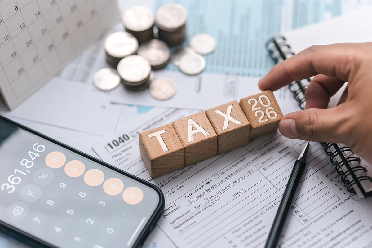 Hand placing a 2026 block beside TAX on tax forms, with a calculator and coins