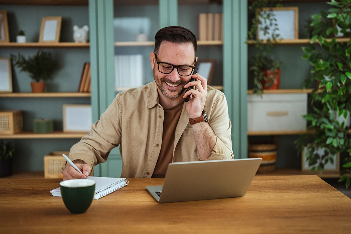 Man speaking on the phone while taking handwritten notes at his laptop about how to start investing in Ireland.