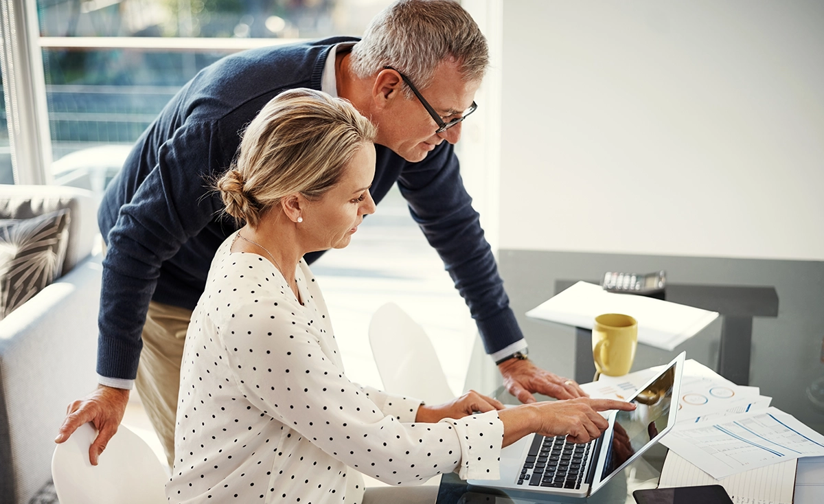A middle aged couple organising their taxes on investments at home on their laptop with paperwork on the table.